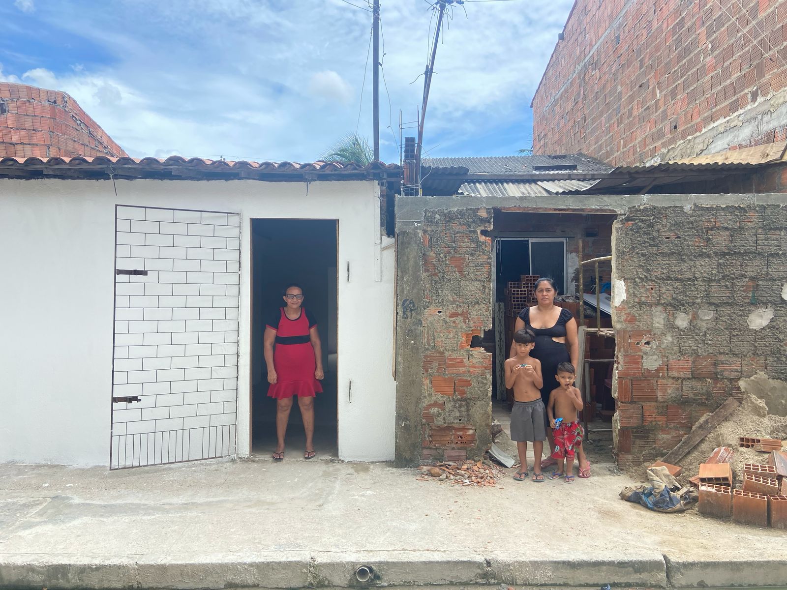 dona vania em frente à sua casa recém pintada de branco e ana paula em frente  à sua casa que ainda está só com os tijolos cinza, sem acabamento